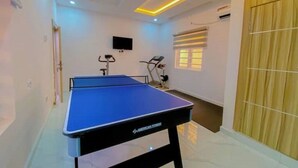 Ein Raum mit einem blauen Tischtennis-Tisch, Fitnessgeräten, einem Fernseher und einem Fenster mit Jalousien.