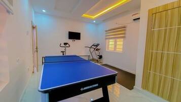 Une pièce avec une table de ping-pong bleue, du matériel de sport, un téléviseur et une fenêtre avec des stores.