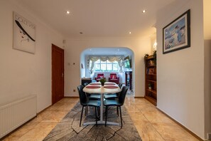 A dining area with a round table, chairs, and a rug. There is a door leading to another room, a bookshelf, and a framed picture on the wall. - 5-bed House - Sleeps 10 - Garden - Parking (Barnet)