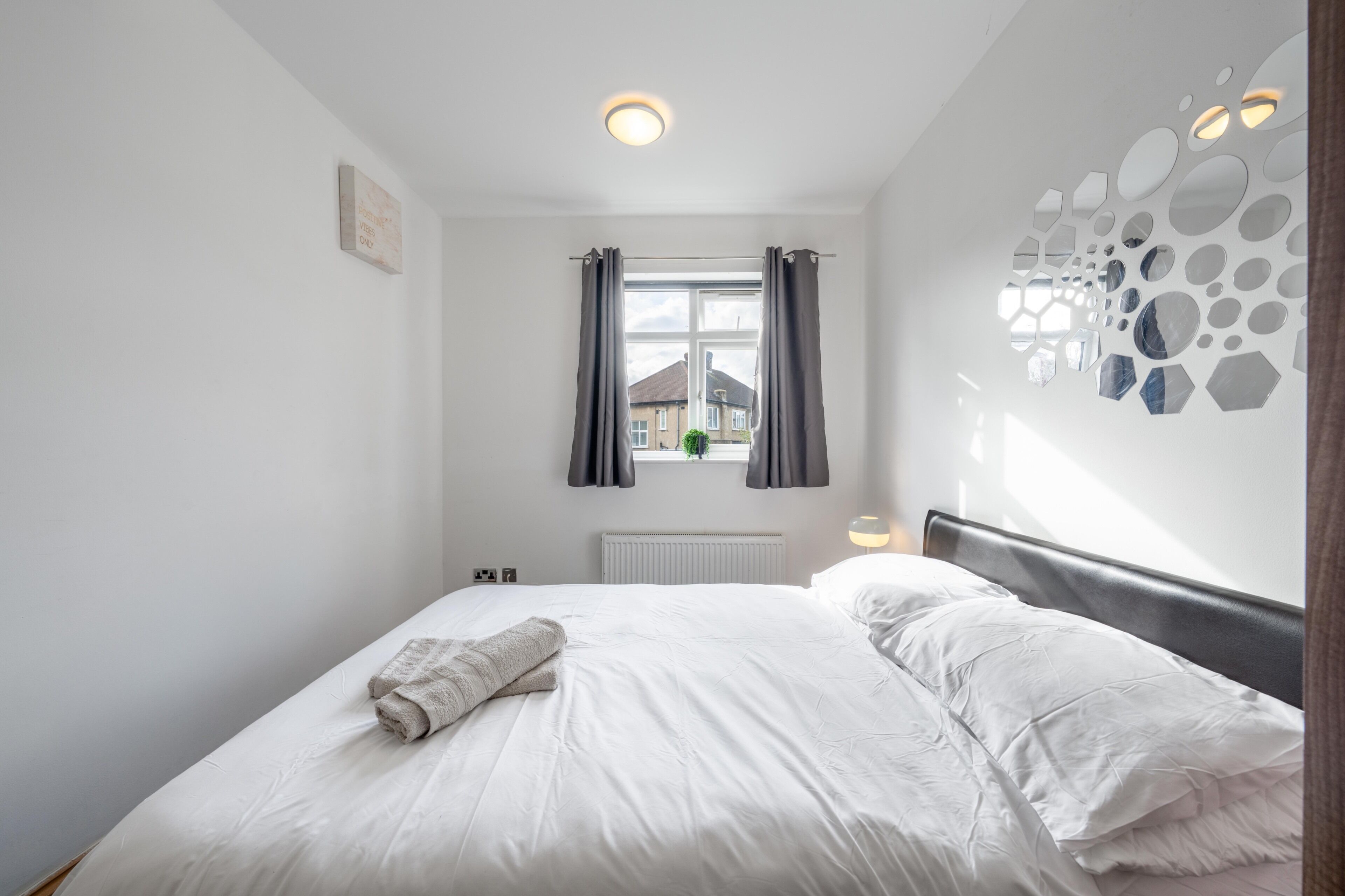 Un letto rifatto con cura, un asciugamano arrotolato, una finestra con tende e un muro decorato con un motivo specchiato geometrico.