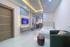 A modern hotel room with a large bed, a TV mounted on the wall, a small seating area, and a door.