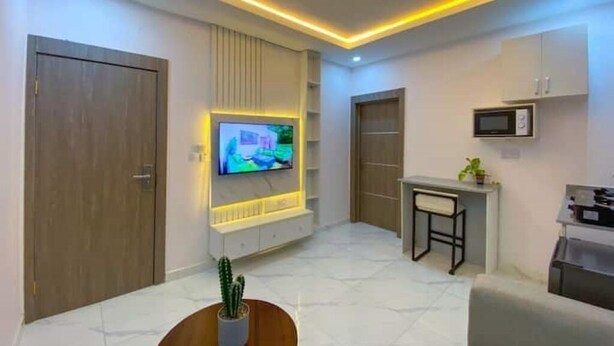 A modern living room with a flat-screen TV mounted on a white wall, a small dining table with a chair, and a kitchen area with a microwave. - 1Bed&Parlour(B4)@Lekki PH 1.24/7 Pwr By NgoZiLivin

 (Lekki)