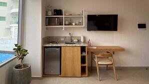 Eine kompakte Küche mit integriertem Kühlschrank, Mikrowelle und Spüle. Es gibt einen Holztisch mit einem Stuhl, einen Blumentopf und einen an der Wand befestigten Fernseher.