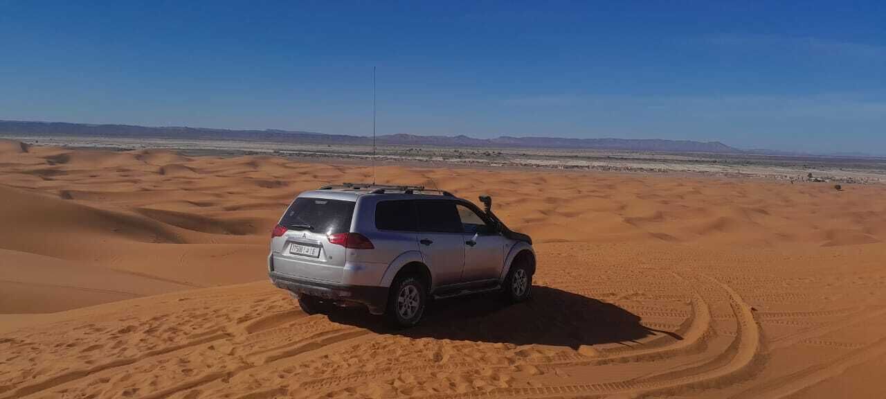 Un SUV plateado estacionado en una duna arenosa bajo un cielo despejado y montañas distantes.