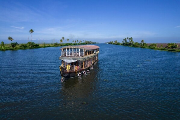 Una casa flotante con cubierta superior protegida y terraza inferior abierta, navegando en un río tranquilo.