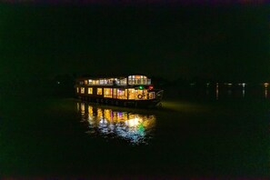 Una amplia casa flotante iluminada con cubierta central y varios niveles, flotando sobre aguas tranquilas de noche.