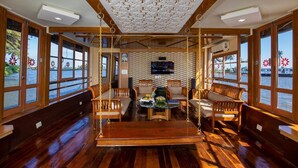 Interior de barco con suelo de madera, ventanas amplias, zona de estar central con sofás y una hamaca, y un televisor de pantalla plana montado en la pared.