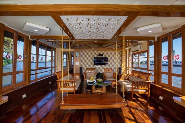 A boat interior with wooden flooring, large windows, a central seating area with sofas and a swing, and a flat-screen TV mounted on the wall.