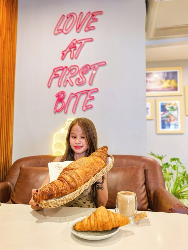 Une personne tenant un grand pain à la main, assise à une table devant une assiette de croissants et une tasse de café.