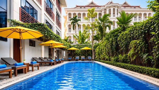 A swimming pool with lounge chairs and umbrellas, surrounded by lush greenery and a large building with classical architecture in the background. - Angkor Mansion & Residence (Siem Reap)