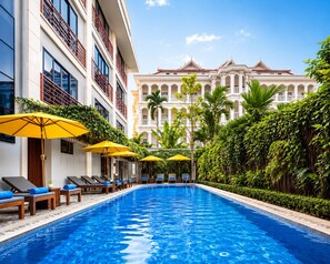 Une piscine entourée de chaises longues et de parasols, bordée d’une végétation luxuriante et avec, en arrière-plan, un grand bâtiment à l’architecture classique.