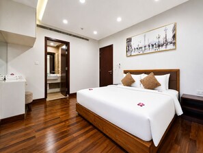 Ein Hotelzimmer mit Holzboden, einem Bett mit weißer Bettwäsche und zwei Kissen, einem Nachttisch, einem Spiegel, einem Badezimmer mit Waschbecken und Spiegel sowie einem gerahmten Bild an der Wand.