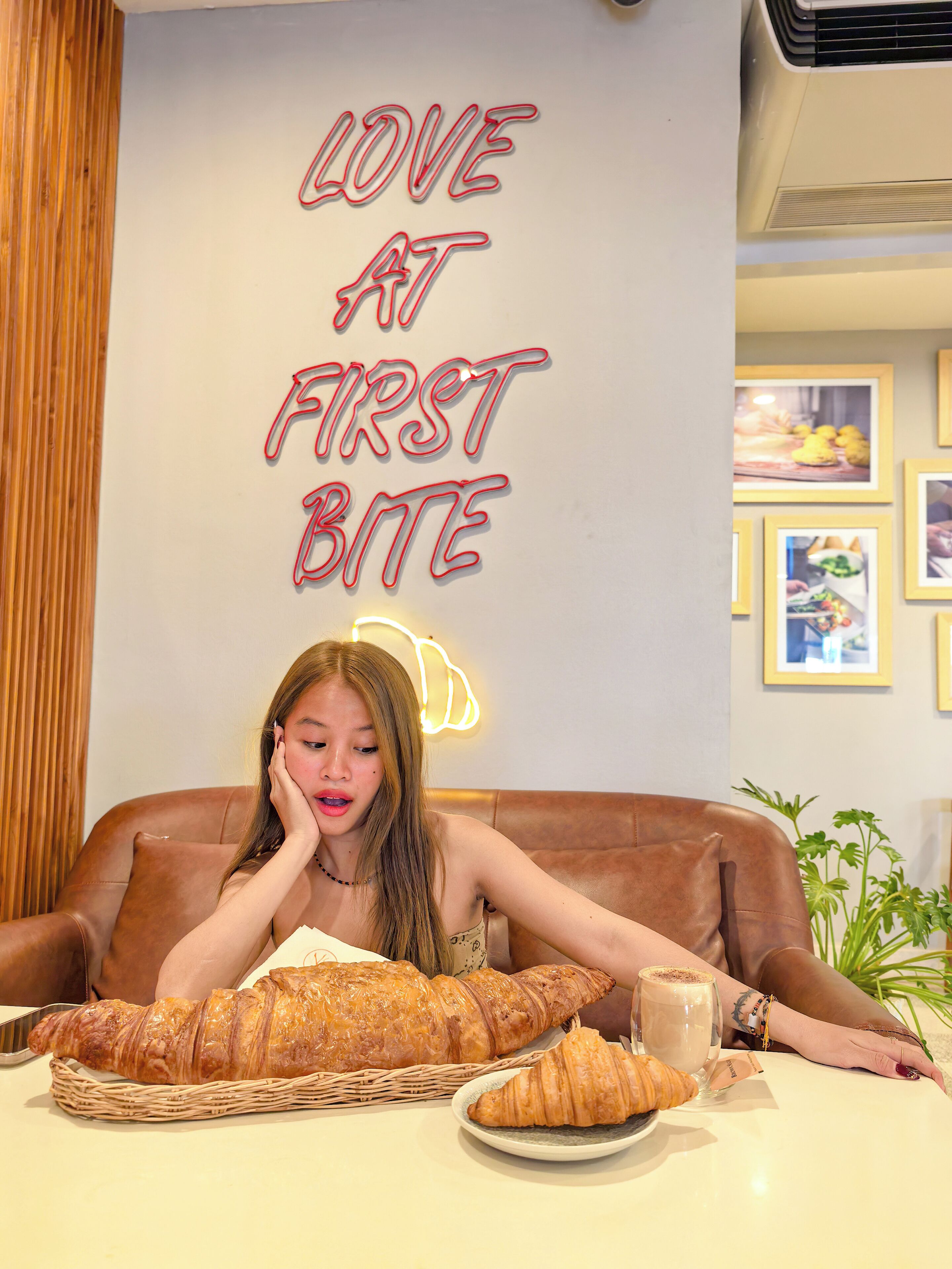 A person seated at a table with a large loaf of bread, a croissant, and a cup of coffee. The wall behind has a neon sign that reads "LOVE AT FIRST BITE."