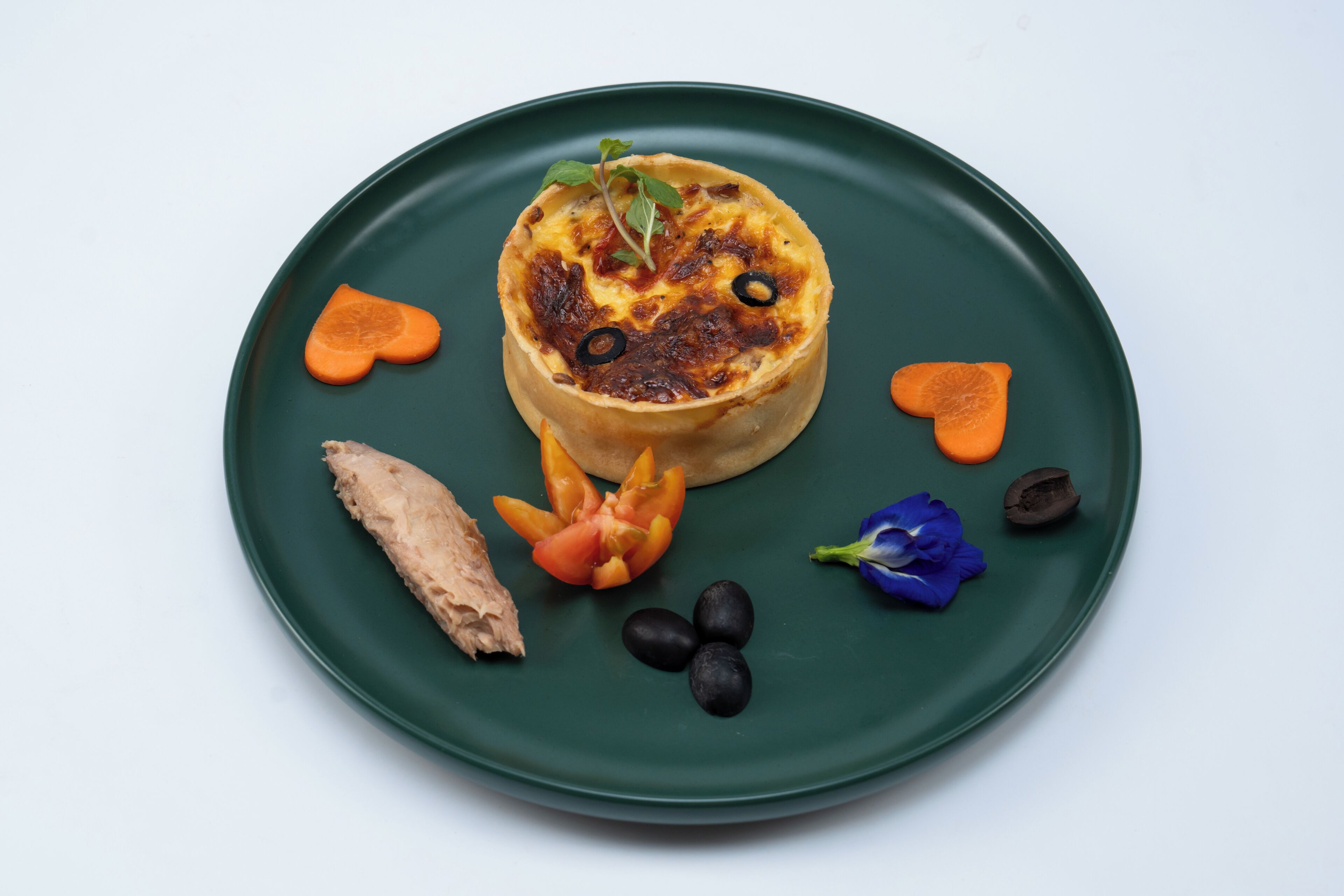 Un plato con una quiche, zanahorias, un tomate, aceitunas y una flor.