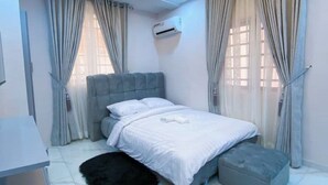 A modern bedroom with a grey upholstered bed, white bedding, and a black rug. - 1Bed&Parlour(B3)@Lekki PH 1.24/7 Pwr By NgoZiLivin

 (Lekki)