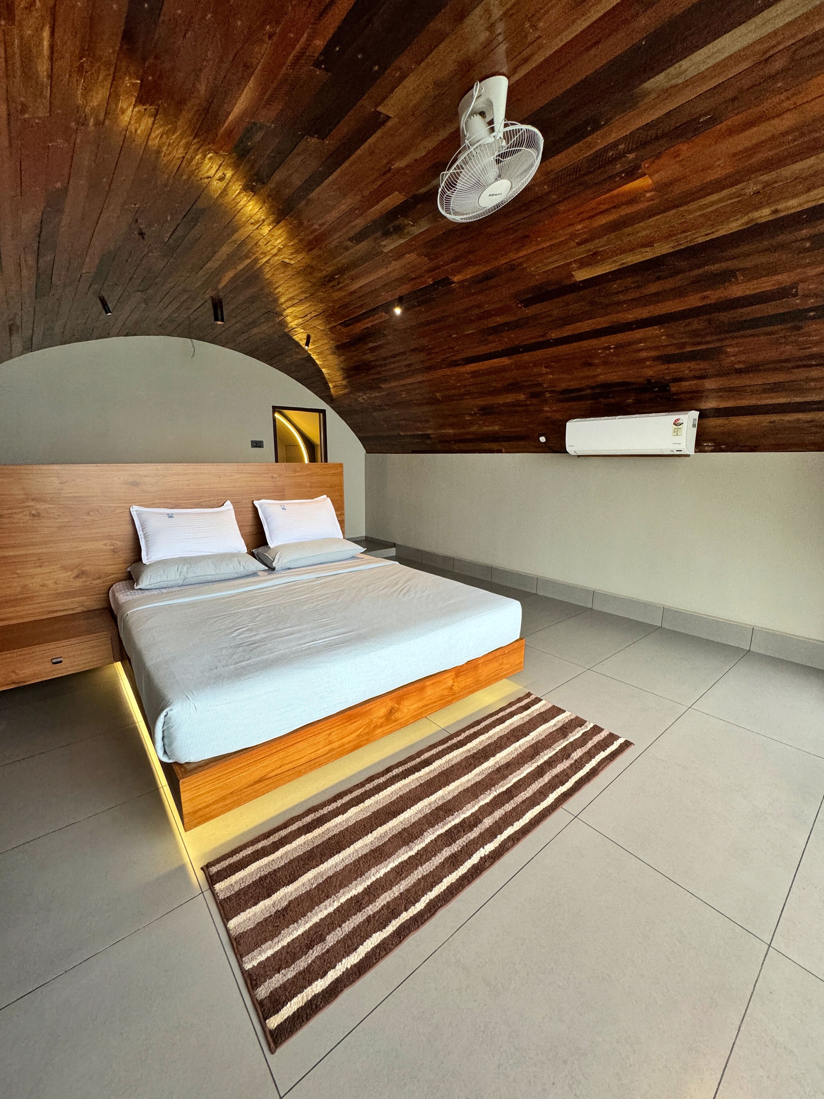 A modern bedroom with a wooden ceiling, a bed with white linens, a standing fan, and a wall-mounted air conditioner.