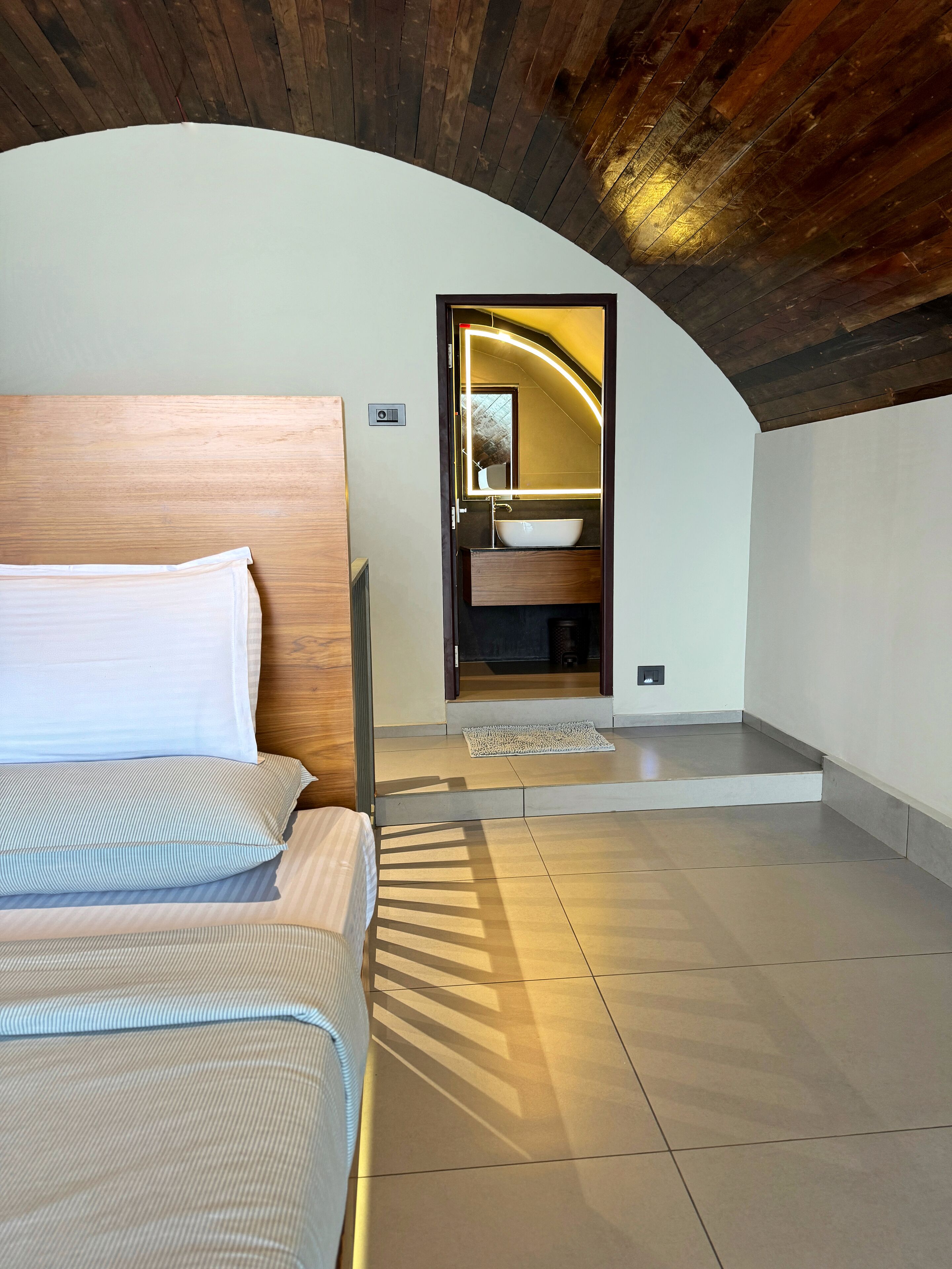 A bedroom with a bed, a wooden headboard, a bathroom with a sink and mirror, and a sloped ceiling.