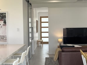 A modern living room with a flat-screen TV, a brown leather sofa, and a dining area with white chairs.