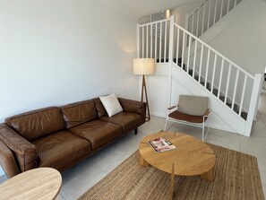 A living room with a brown leather sofa, a wooden coffee table, a white armchair, and a staircase with white railings.