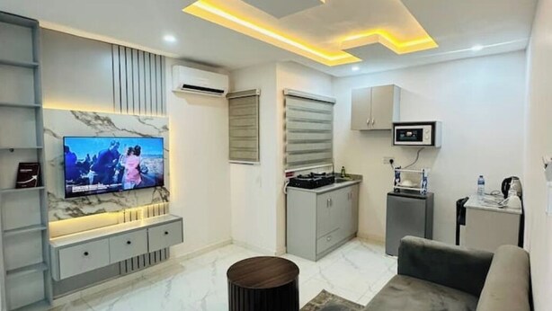 A modern living room with a flat-screen TV, a built-in entertainment unit, a kitchen area, and a seating area with a sofa and ottoman. - 1Bed&Parlour(B2) @ Lekki PH 1.24/7 Pwr By NgoZiLivin

 (Lekki)