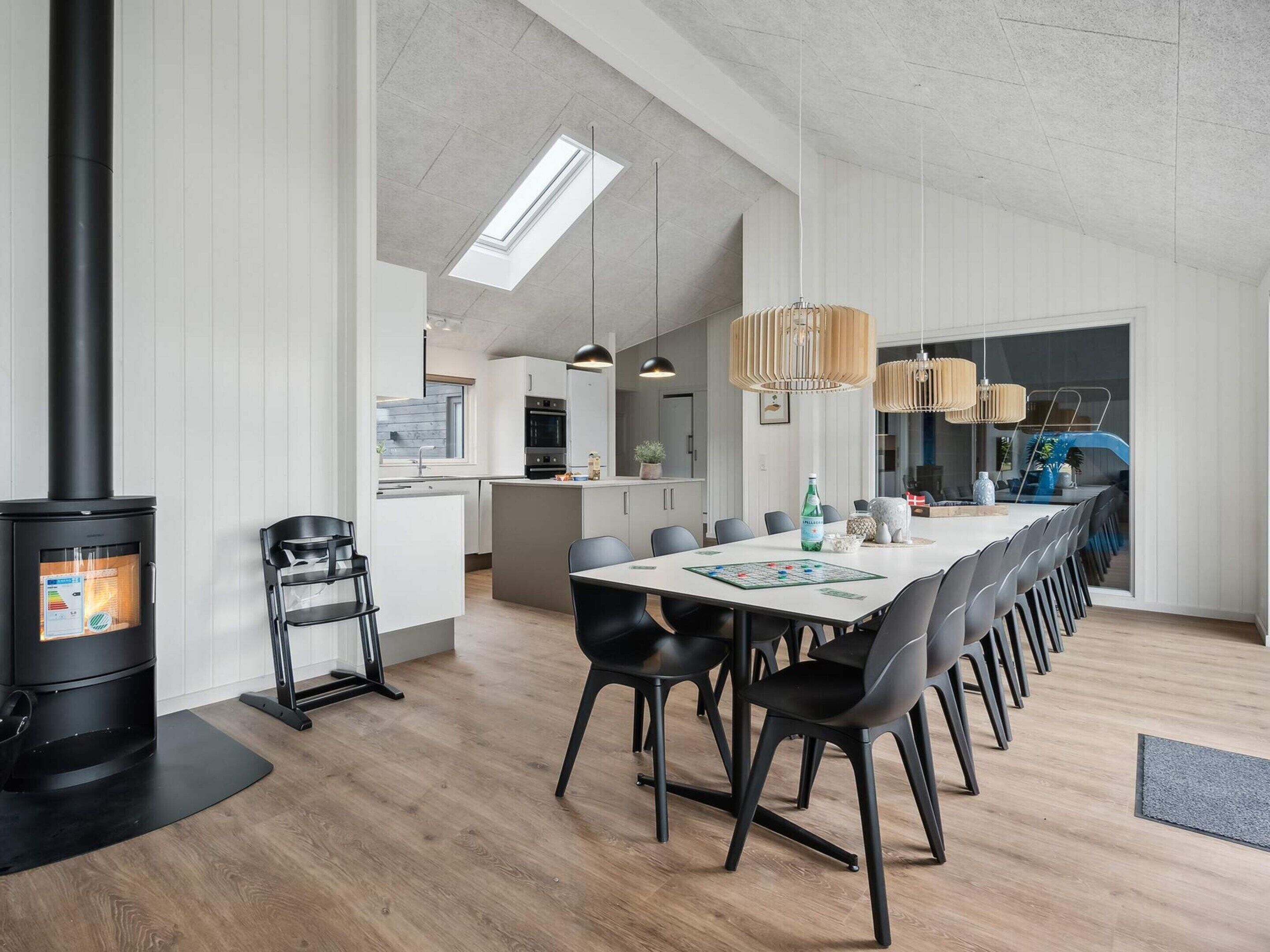 A modern kitchen with a built-in stove, a dining area with a long table and chairs, and a view of the living area through a glass door.