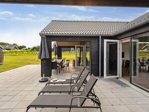 A covered outdoor seating area with chairs and tables, a patio with a tiled floor, and a view of a house in the background.