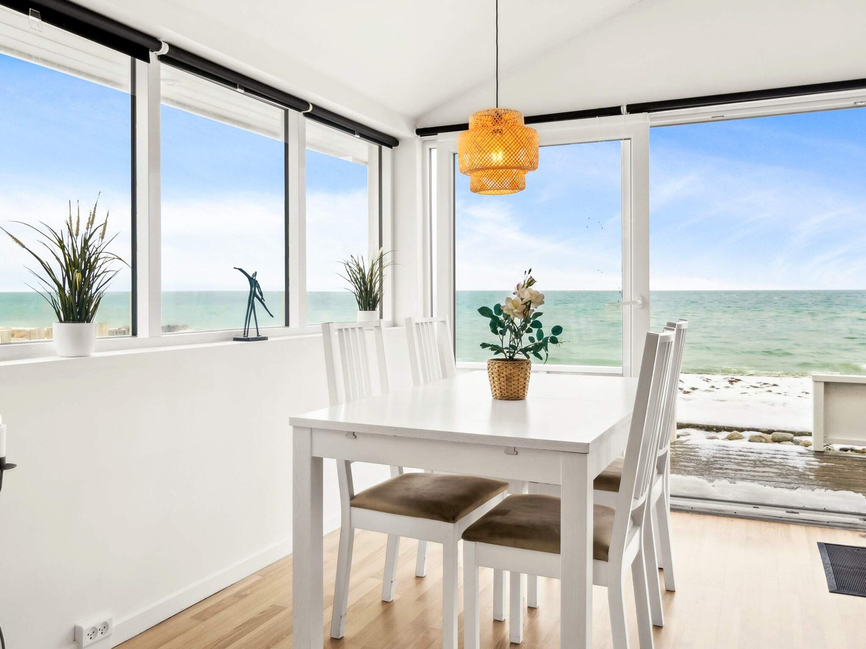 A dining area with a white table and chairs, large windows overlooking the sea, and a hanging light fixture.