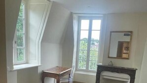 A bedroom with a bed, a desk, and a chair. There is a window providing natural light and a view of greenery outside. - IL MAESTRO - HOTEL RESTAURANT DU CHATEAU (Ecouen)