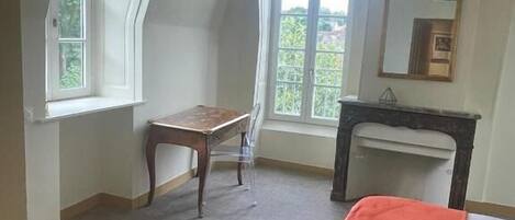 A bedroom with a bed, a desk, and a chair. There is a window providing natural light and a view of greenery outside.