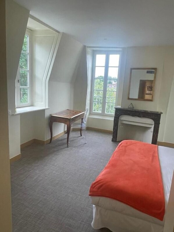 A bedroom with a bed, a desk, and a chair. There is a window providing natural light and a view of greenery outside.