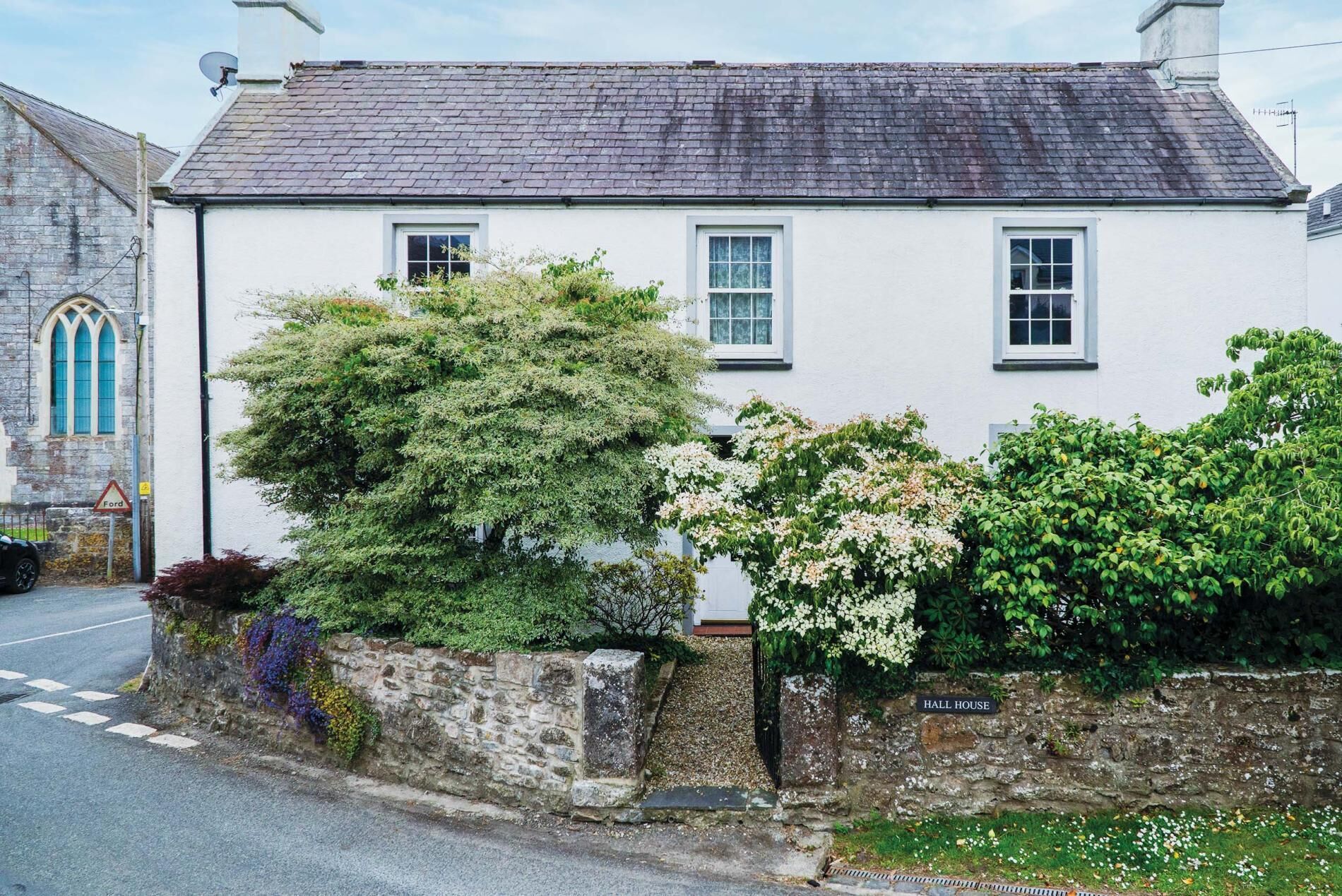 A white building with a stone wall, a sign reading 'HALL HOUSE,' and a road with a 'Ford' sign.