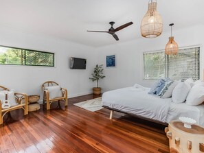 A bedroom with a bed, two wicker chairs, a ceiling fan, and a large window with blinds.