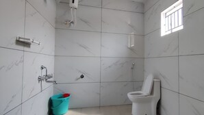 A bathroom with white marble tiles, a toilet, a shower, and a bucket. - The Hill View Guesthouse (Kumta)