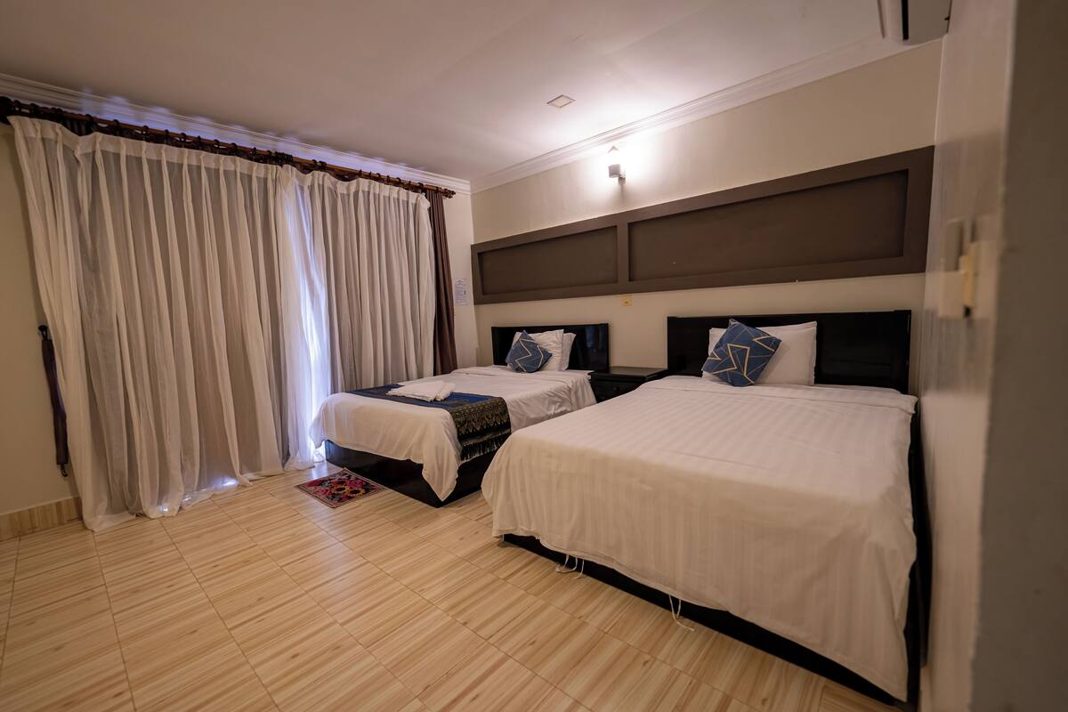 A hotel room with two beds, a large window with sheer curtains, a wooden floor, and a modern headboard.