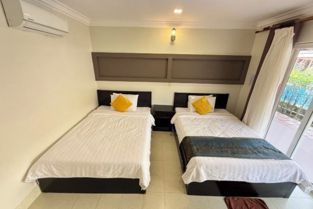Habitación de hotel con dos camas, una mesita de noche, una lámpara de pared y vistas al exterior.