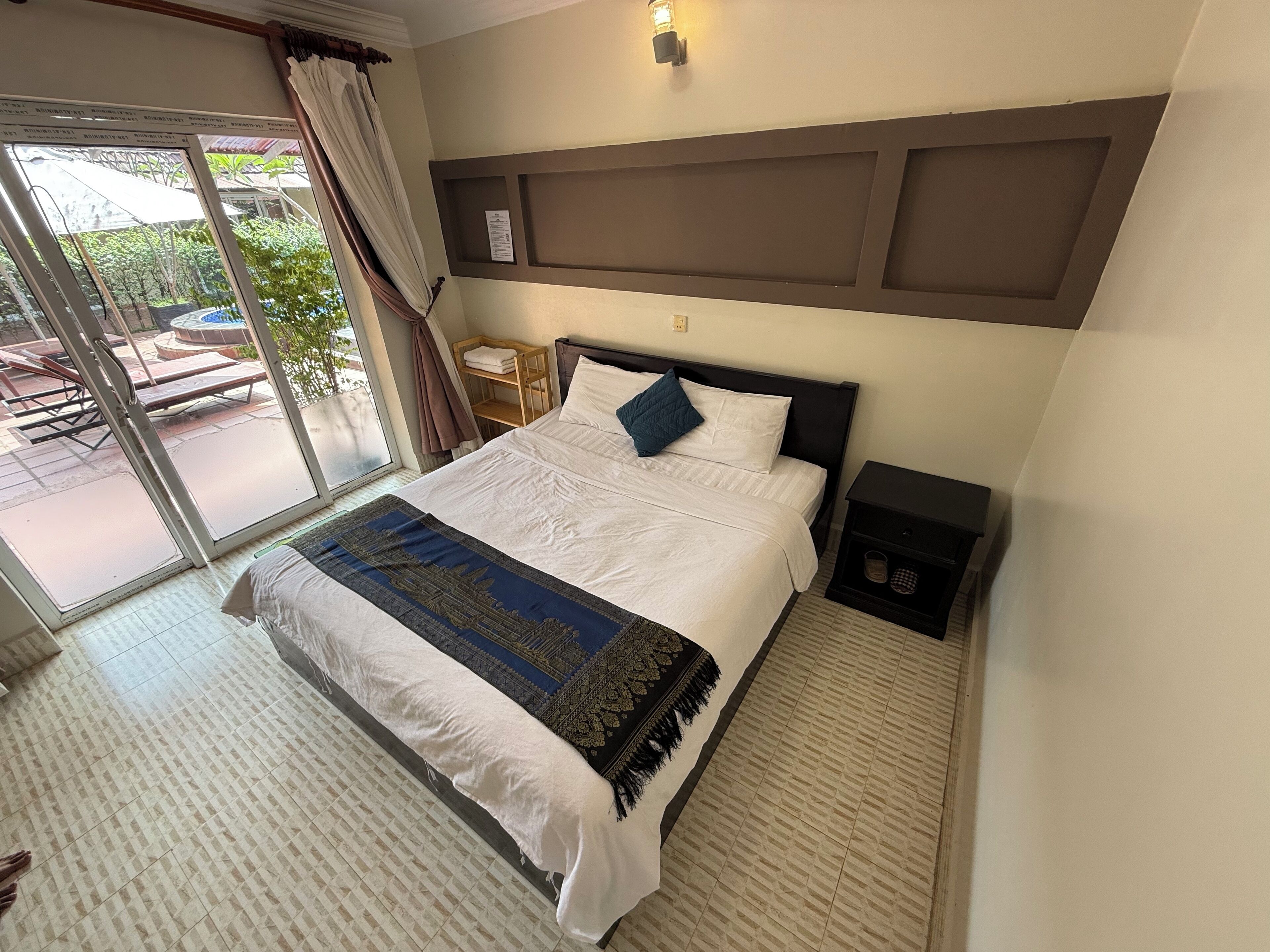 A hotel room with a bed, a nightstand, a wall-mounted shelf, and a sliding glass door leading to an outdoor area.