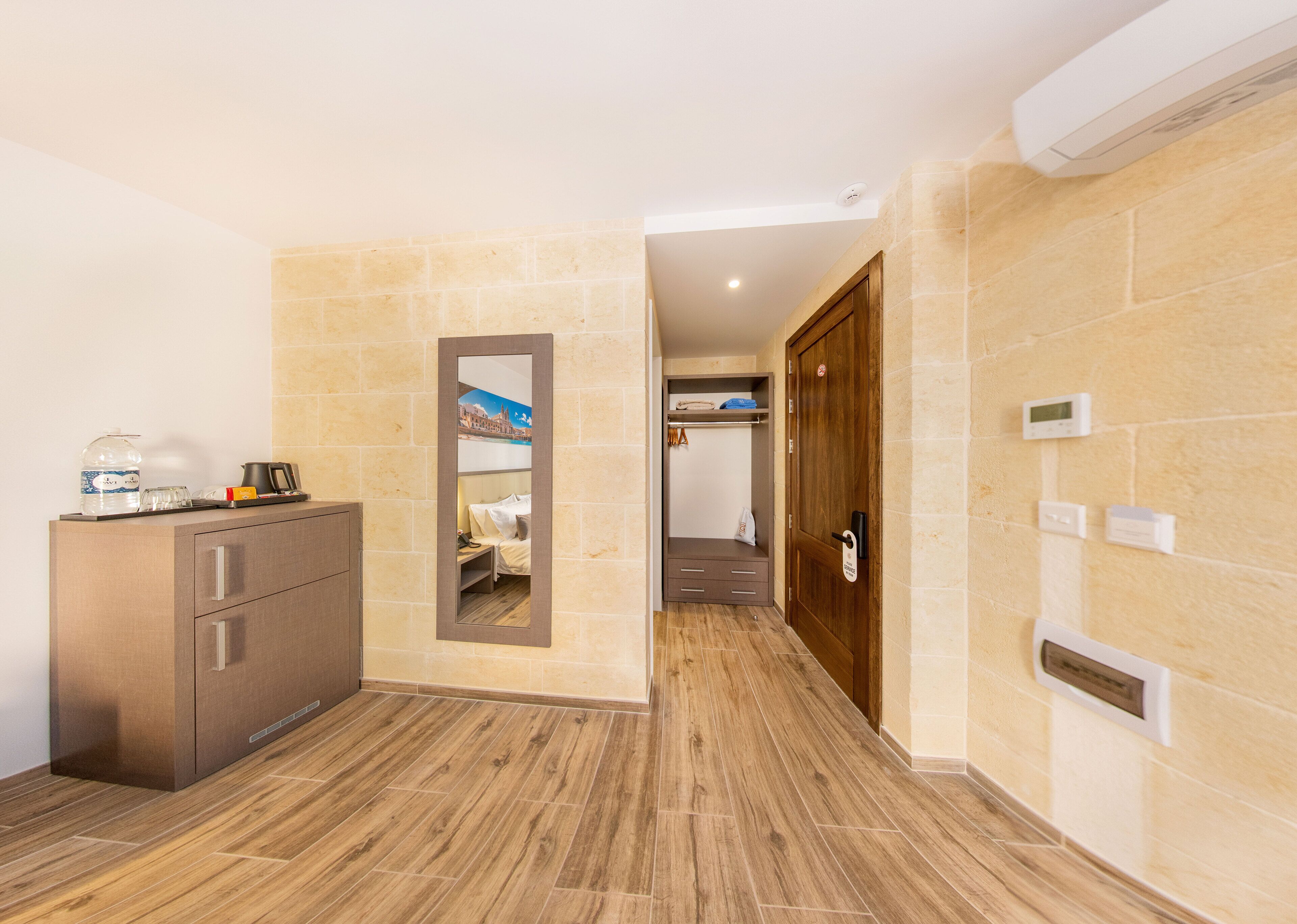 Quarto de hotel com um armário de madeira, um espelho, uma cama com cabeceira, um guarda-roupa e uma porta com puxador.