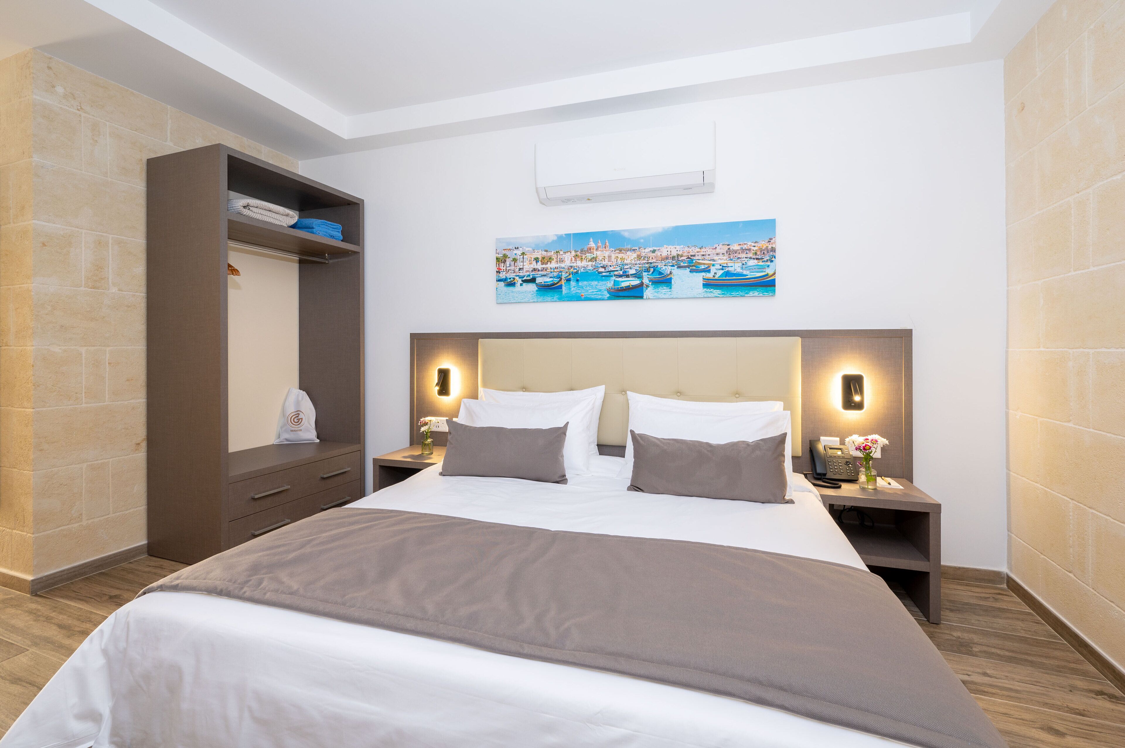 Habitación de hotel con cama, mesita de noche, aire acondicionado mural y un cuadro de un pueblo costero, además de un armario.