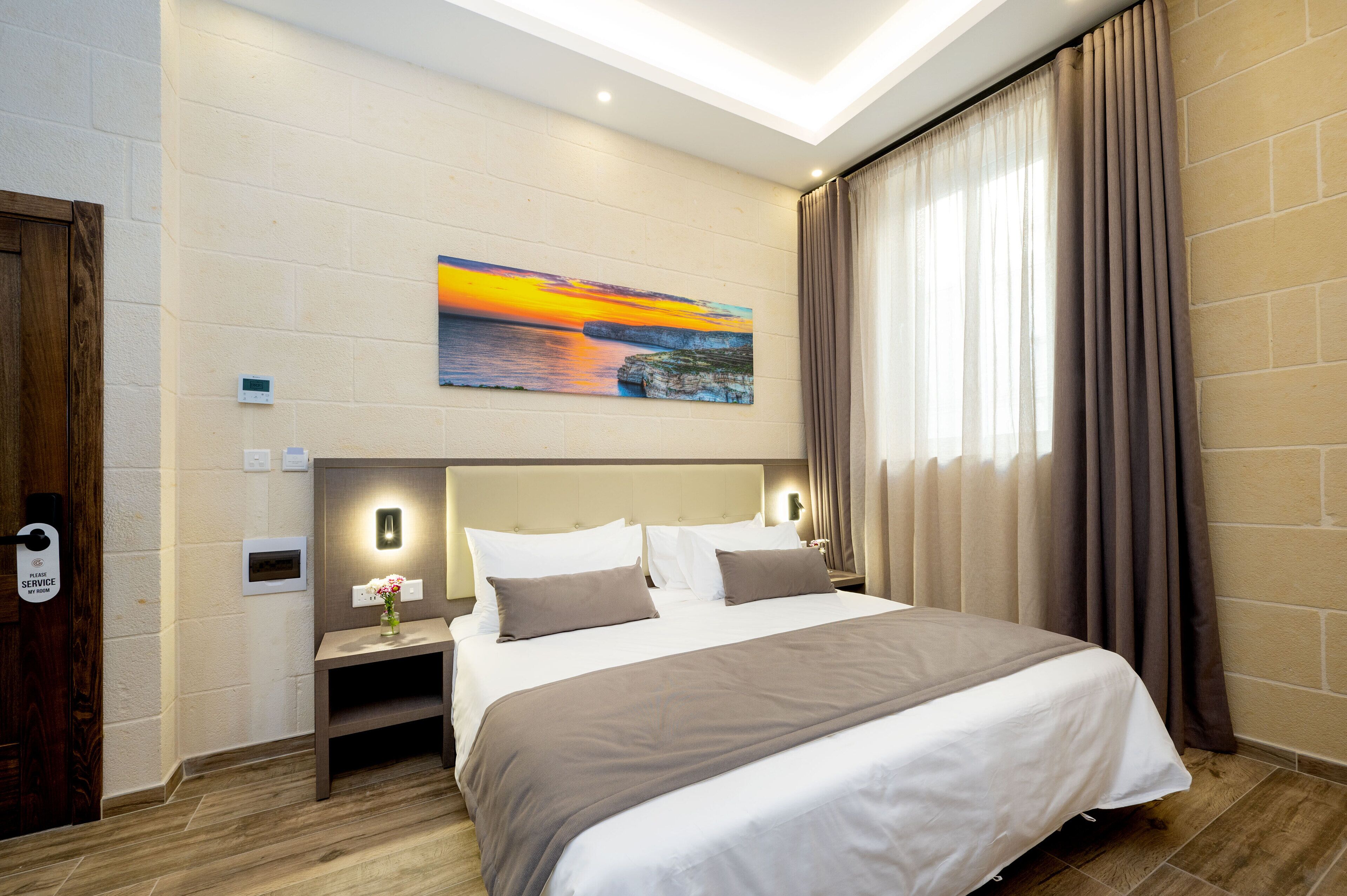 A hotel room with a bed, a nightstand, a painting, a window with curtains, and a door with a service sign.