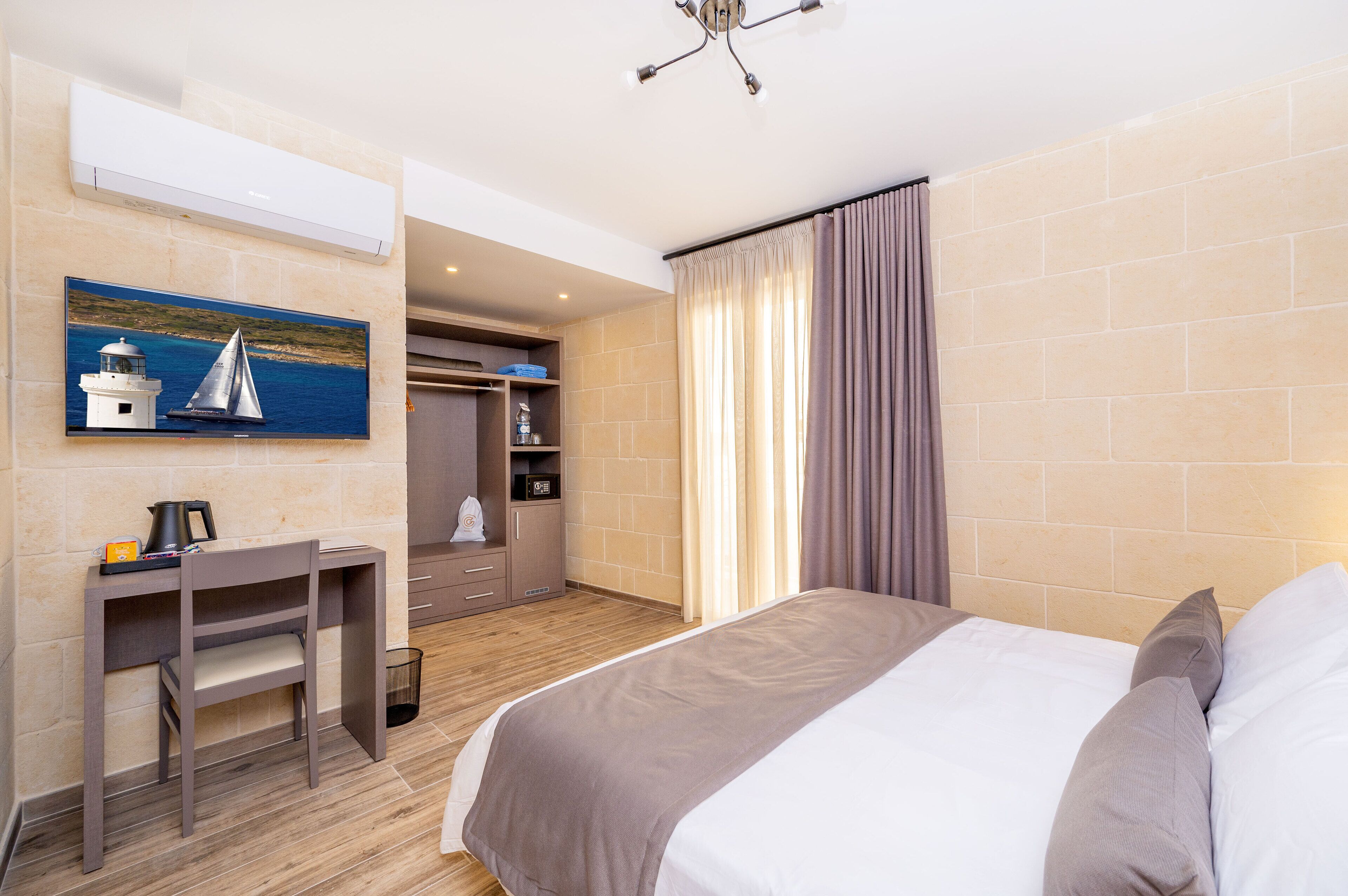 Une chambre d’hôtel comprenant un lit, un bureau, une chaise, un tableau représentant un phare et un voilier, ainsi qu’une armoire.