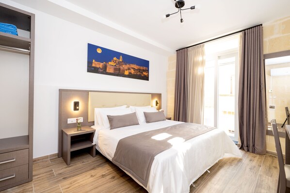 A modern hotel room with a large bed, a nightstand, a wall-mounted light, and a painting of a cityscape. - C4 Retreats Gozo (Xaghra)