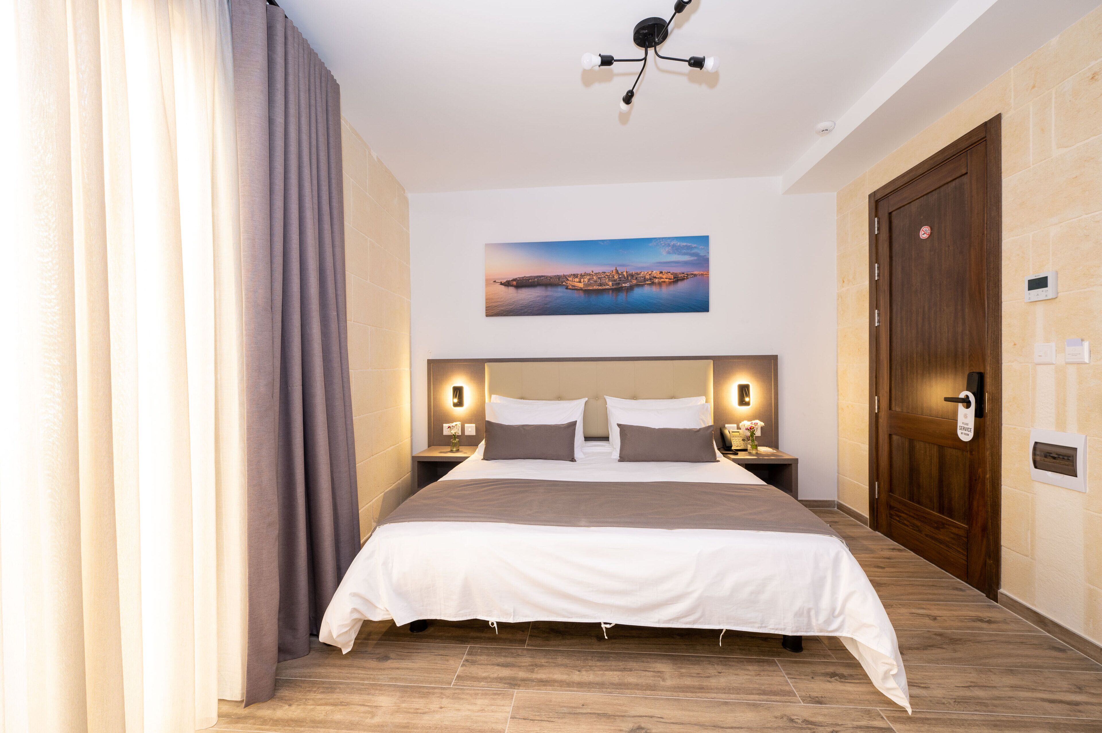 Une chambre d’hôtel avec un grand lit, un plancher en bois, un tableau représentant un paysage urbain accroché au mur et un ventilateur de plafond.
