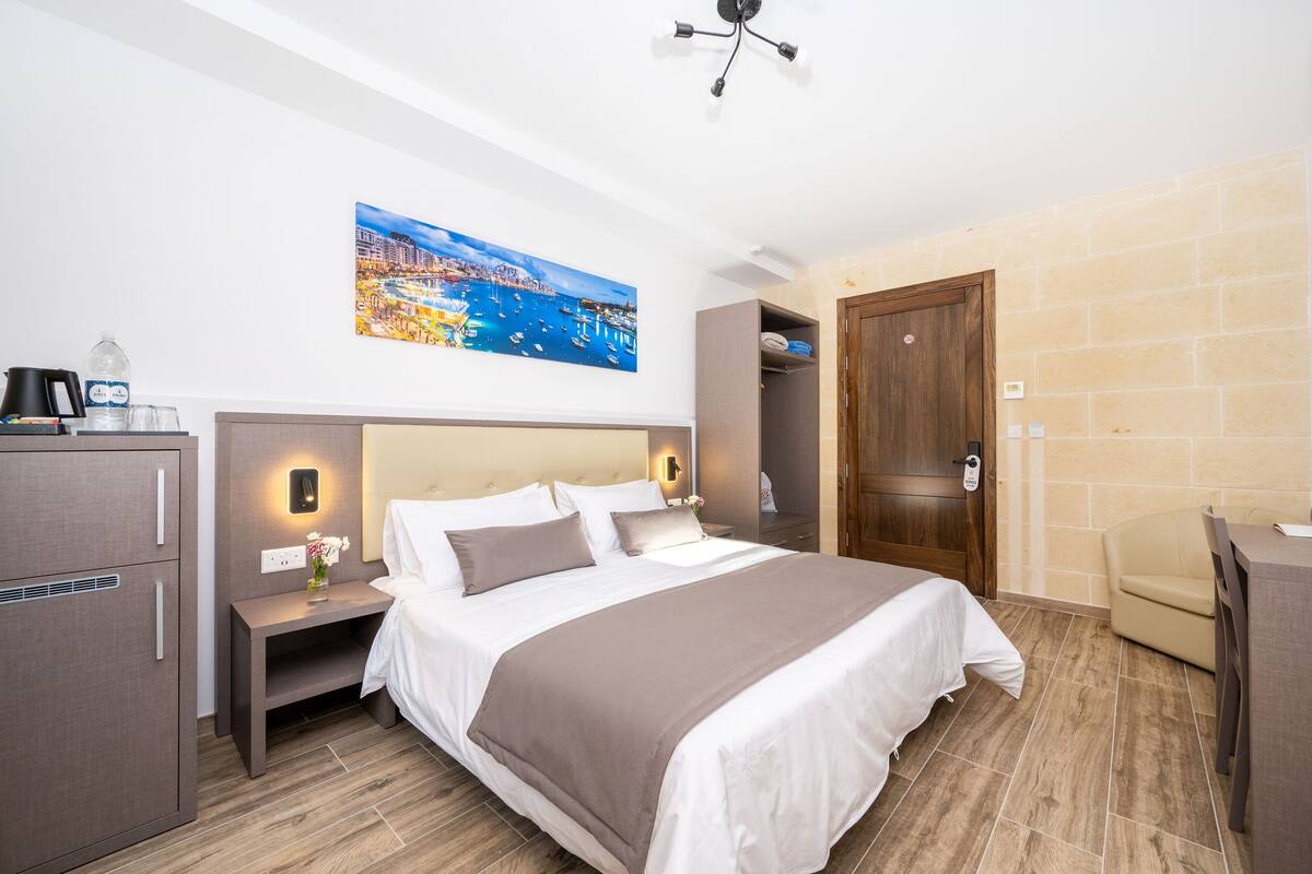 Una habitación de hotel con cama, mesita de noche, lámpara y un cuadro de una ciudad costera.