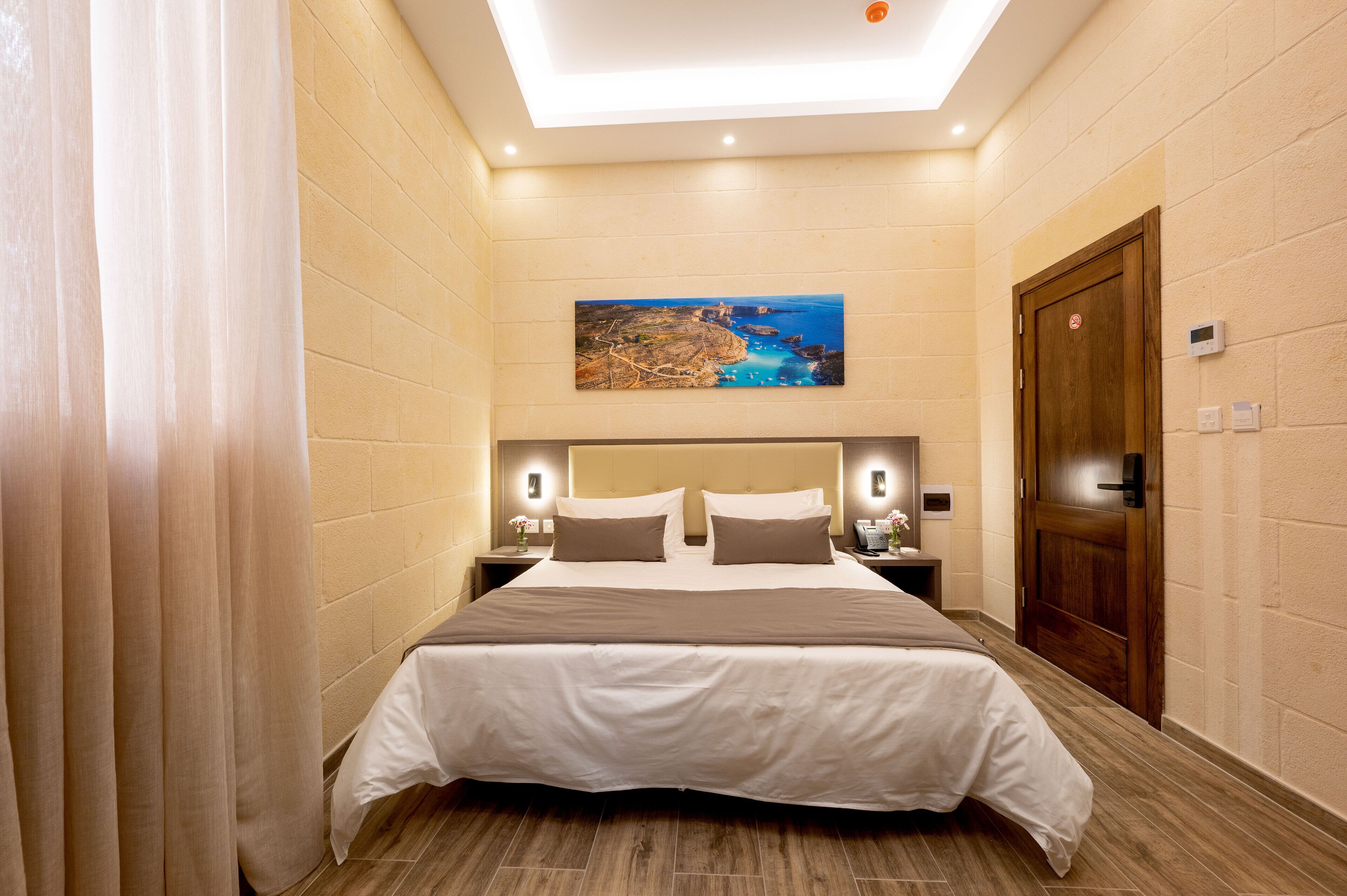 A hotel room with a large bed, a painting of a coastal landscape, a wooden door, and a nightstand with a phone and lamp.