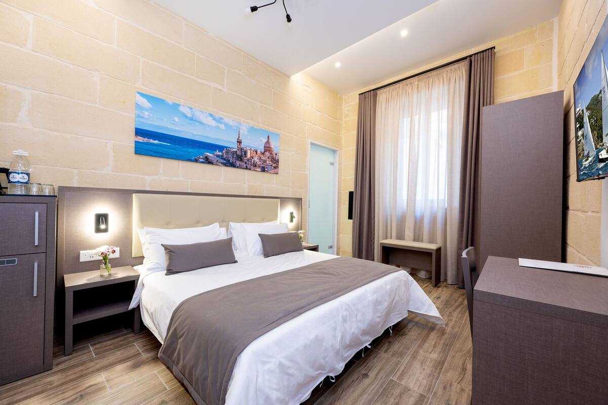 Una habitación de hotel moderna con una cama grande, una mesita de noche con lámpara, un armario y un cuadro de una ciudad costera.