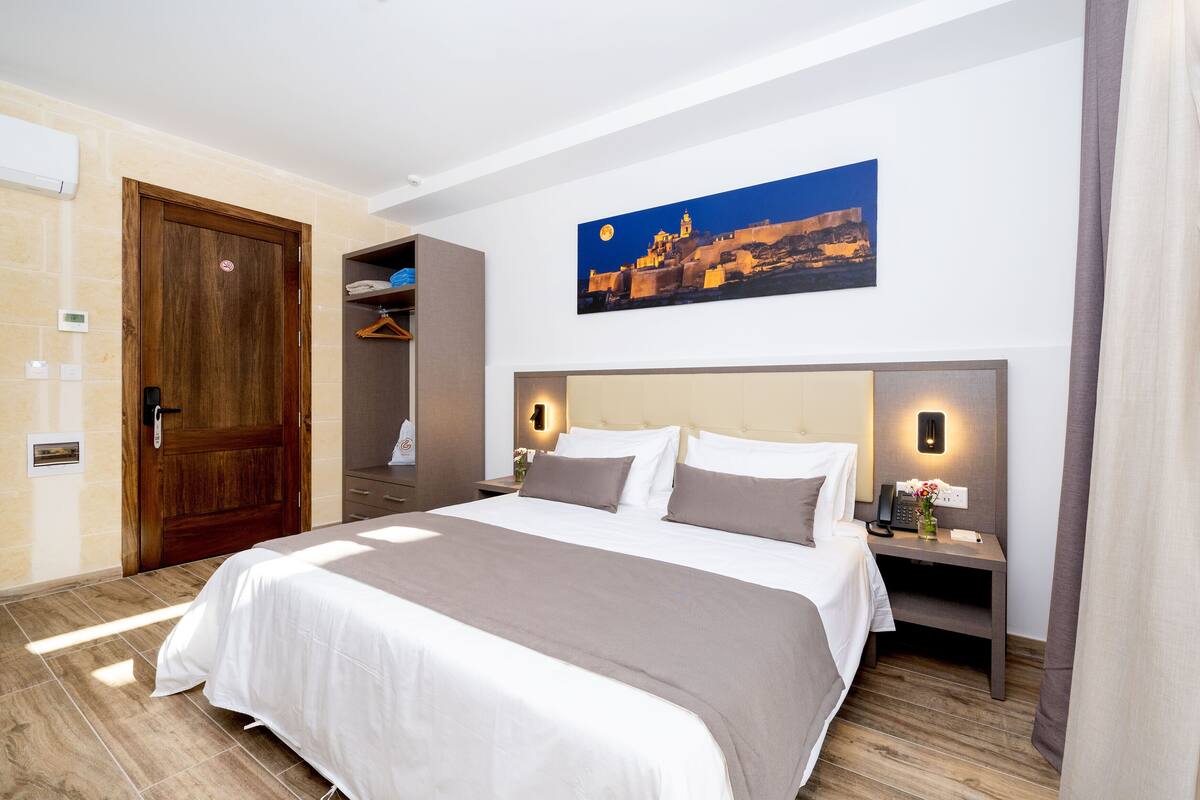 Habitación de hotel con una cama grande, una mesita de noche con teléfono, un cuadro de un paisaje urbano y un armario.