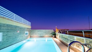 An indoor swimming pool with a wooden wall and a stone wall, a metal railing, and a view of the city skyline at dusk. - C4 Retreats Gozo (Xaghra)