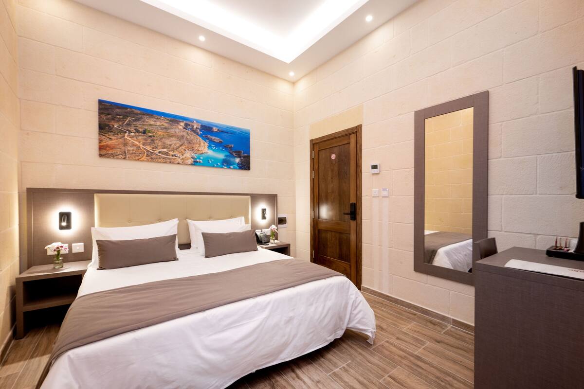 Una habitación de hotel moderna con una cama grande, una mesita de noche con lámpara, una puerta de madera y un espejo amplio.