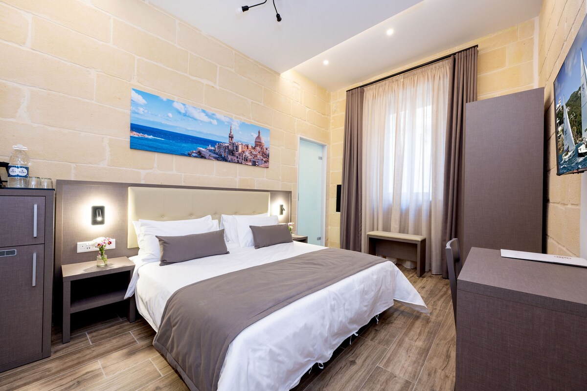 Una habitación de hotel moderna con una cama grande, una mesita de noche con lámpara, un armario y un cuadro de una ciudad costera.