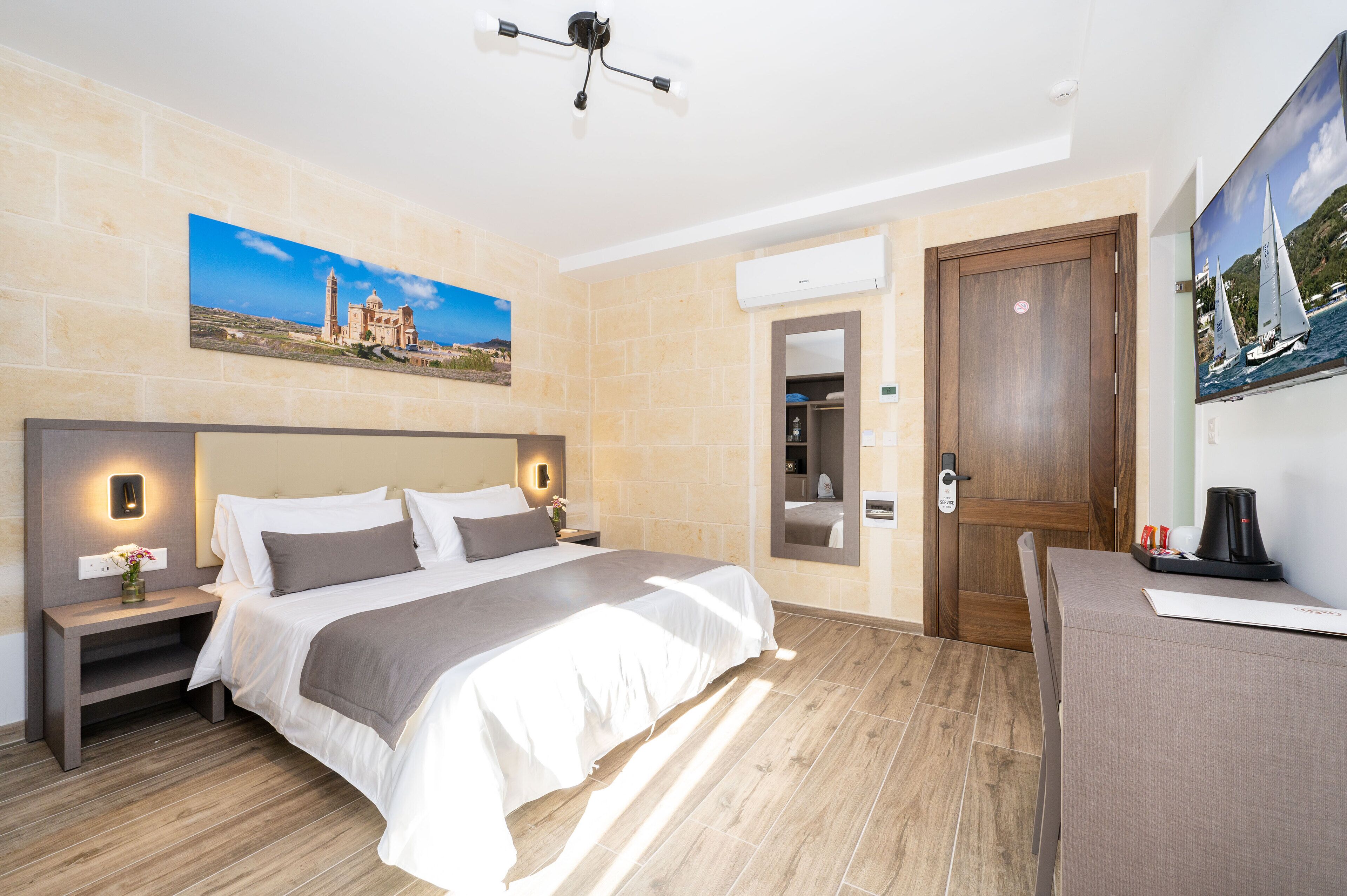 A hotel room with a large bed, a desk, a chair, a nightstand, a wall-mounted TV, and a painting of a coastal scene.